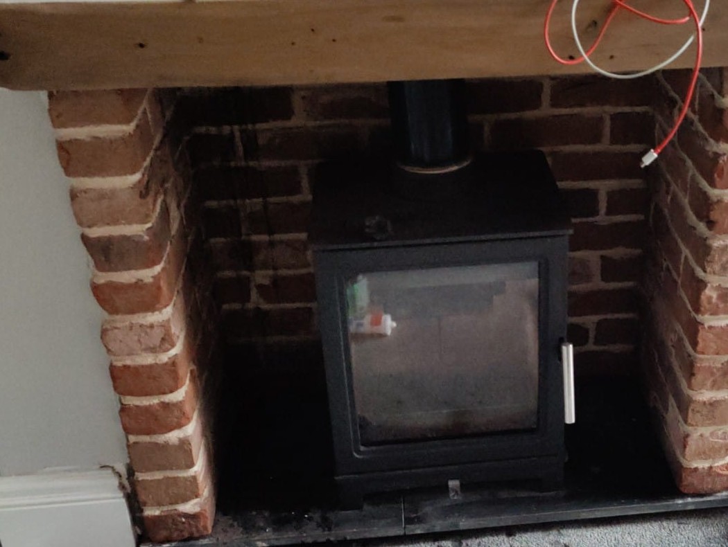 An exposed brick fireplace with an oak beam above a black wood burning stove